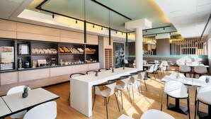 A modern cafe with a central counter displaying pastries and beverages, surrounded by white tables and chairs. - B&B Hotel Salzburg-Nord (Salzburg)