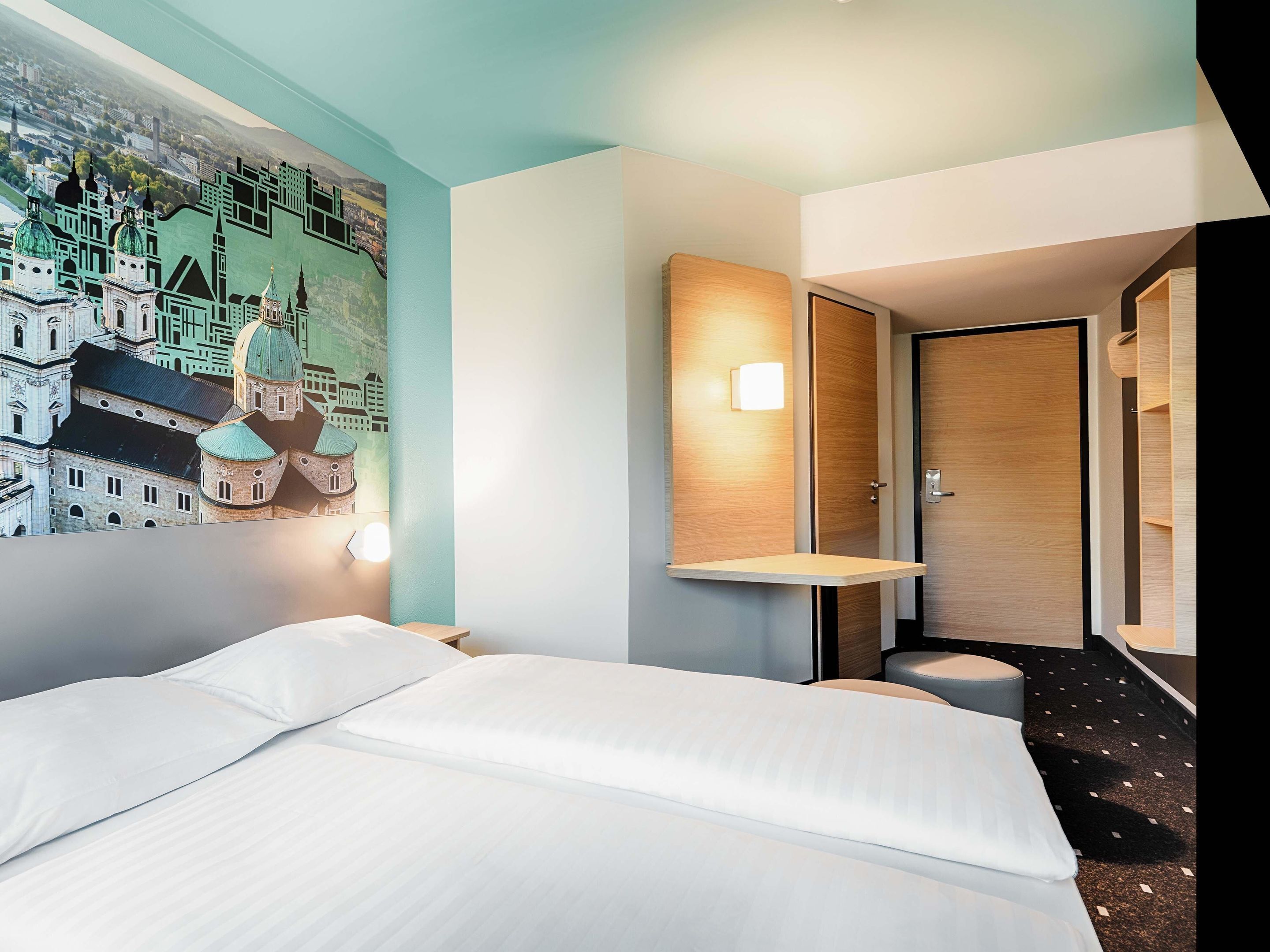 A hotel room with a bed, a nightstand, a wall-mounted lamp, and a door.