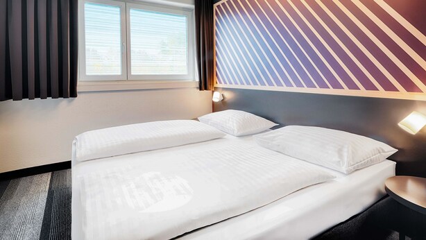 A hotel room with a bed, a window with blinds, a wall with a striped pattern, and a nightstand with a lamp. - B&B Hotel Salzburg-Nord (Salzburg)