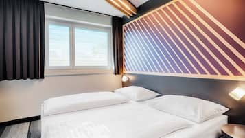 Ein Hotelzimmer mit Bett, Fenster mit Jalousien, einer Wand mit Streifenmuster und einem Nachttisch mit Lampe.