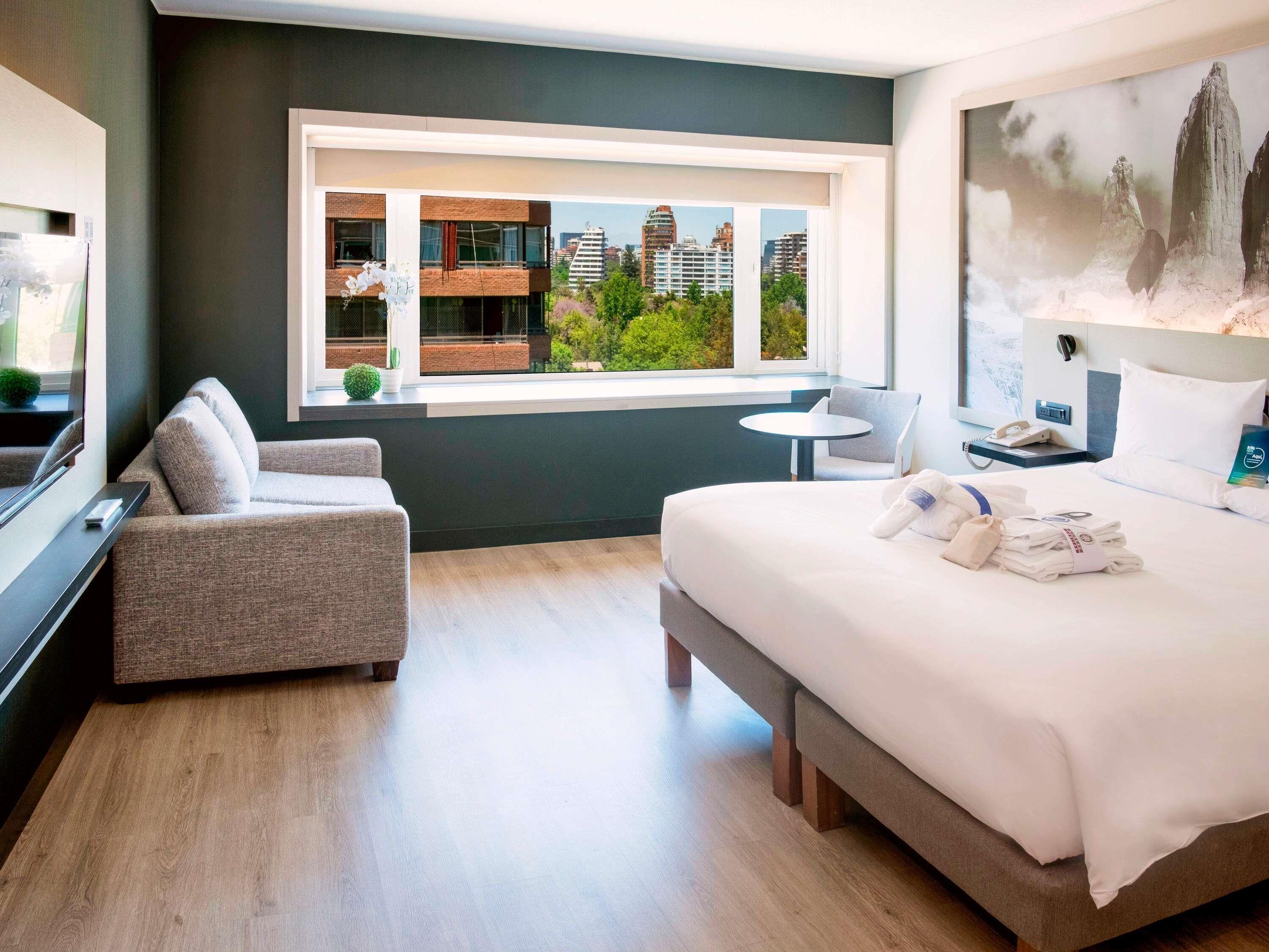 Una habitación de hotel moderna con una cama grande, un sofá pequeño, una mesa redonda y un ventanal con vista a la ciudad.