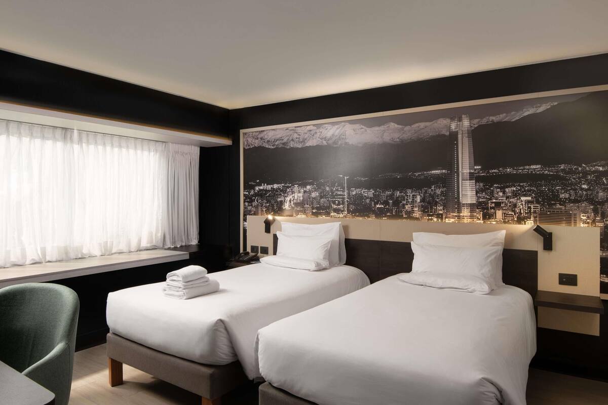 A hotel room with two beds, a cityscape view, a window with curtains, and a green chair.