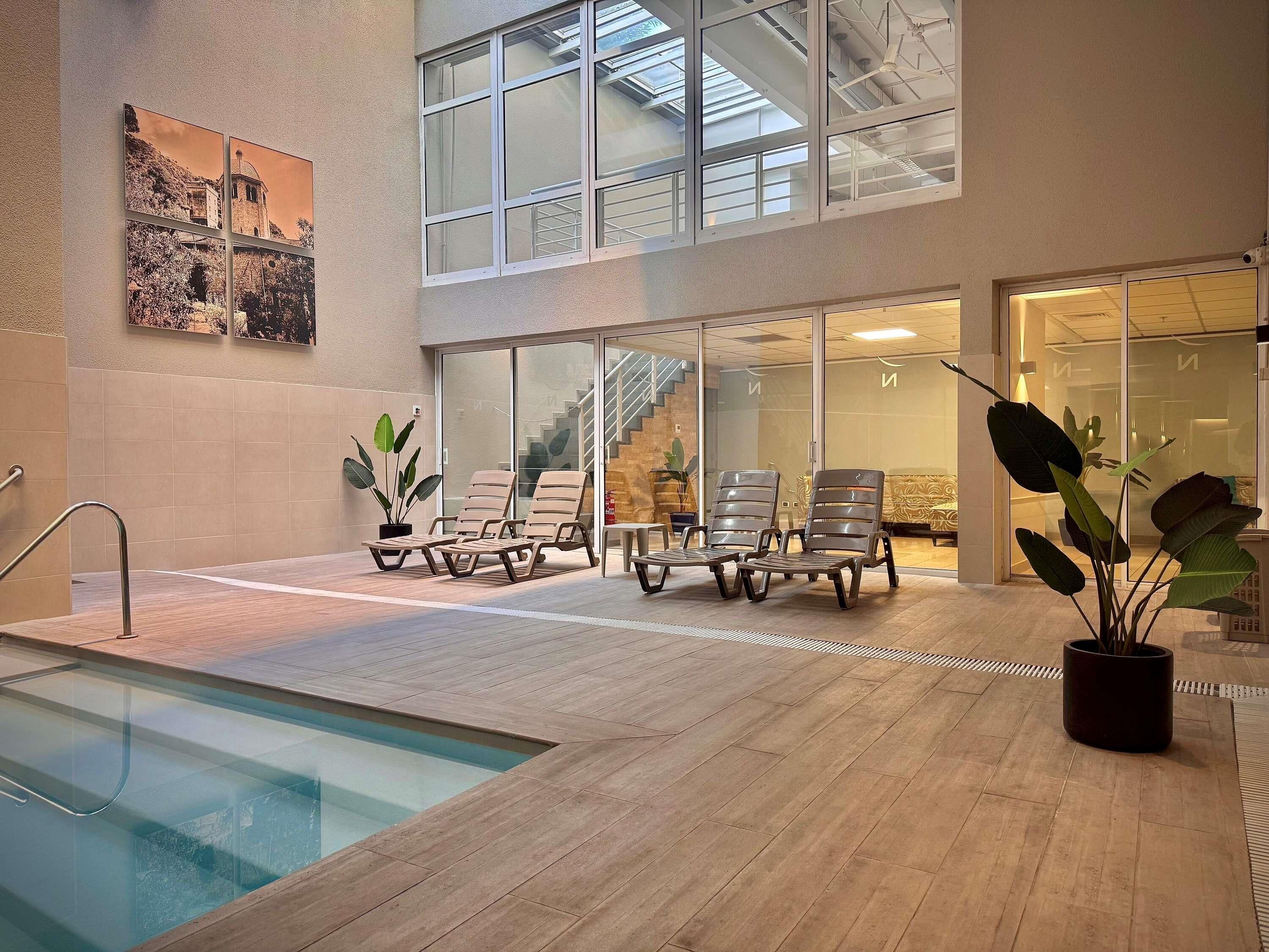 Zona de piscina interior con sillones de descanso, una pared de vidrio y una escalera que lleva a un nivel superior.