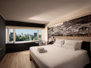 A bedroom with a bed, a window with a city view, a small table, and a wall-mounted phone. - Hotel Novotel Santiago Vitacura (Santiago)