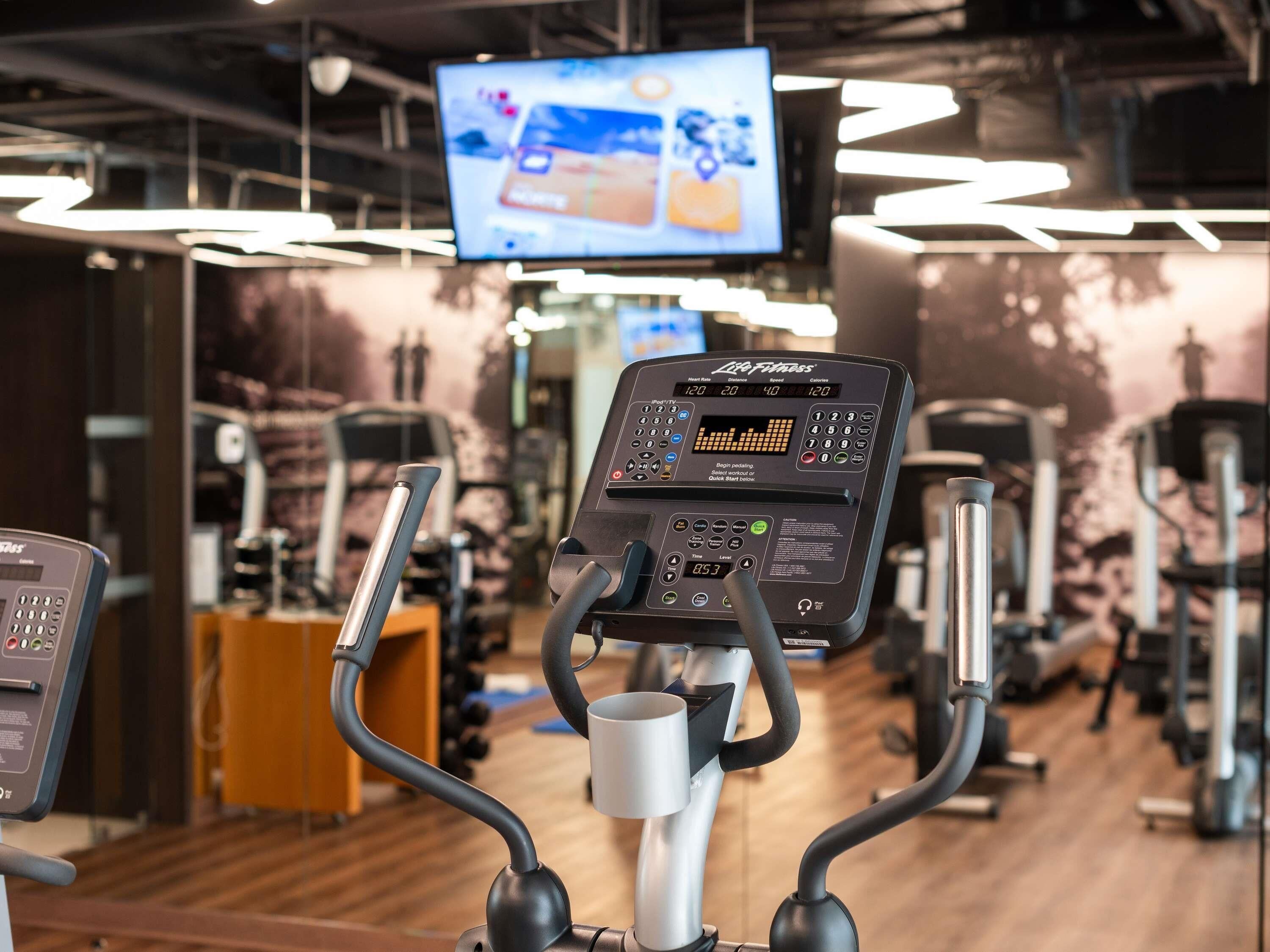 Un appareil d’exercice doté d’un écran numérique, avec un téléviseur placé au-dessus, et divers équipements de salle de sport en arrière-plan.