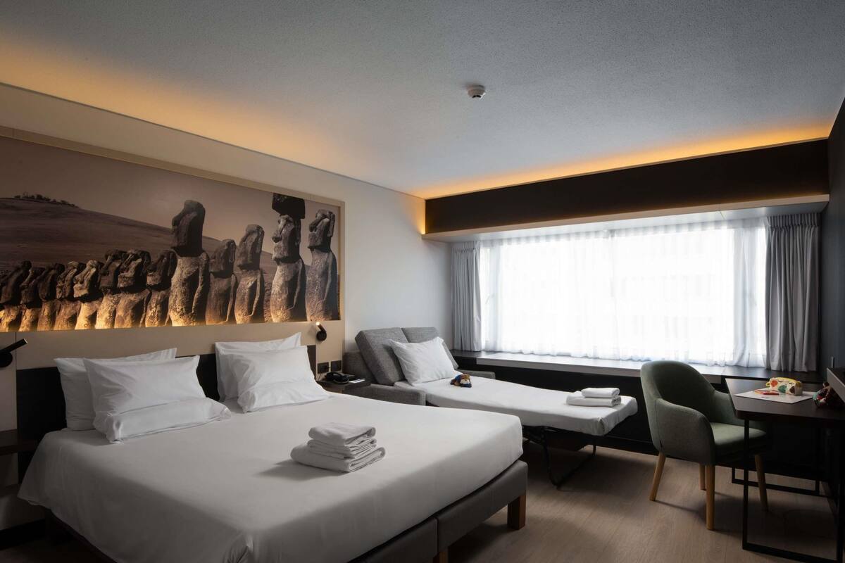 A hotel room with two beds, a desk, a chair, a large window with curtains, and a wall-mounted artwork of Moai statues.