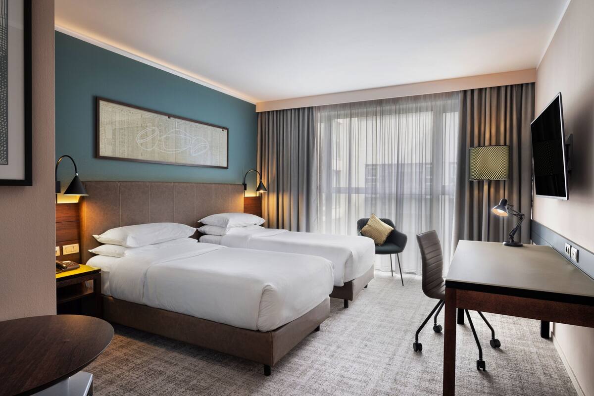 A hotel room with two beds, a desk, a chair, a TV, and a large window with curtains.