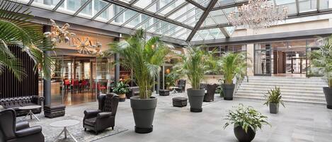 A spacious atrium with a high ceiling, large potted plants, leather seating, and a modern chandelier.