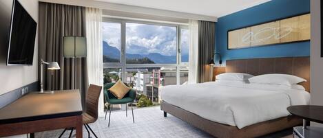A hotel room with a bed, desk, chair, and a large window offering a city view.