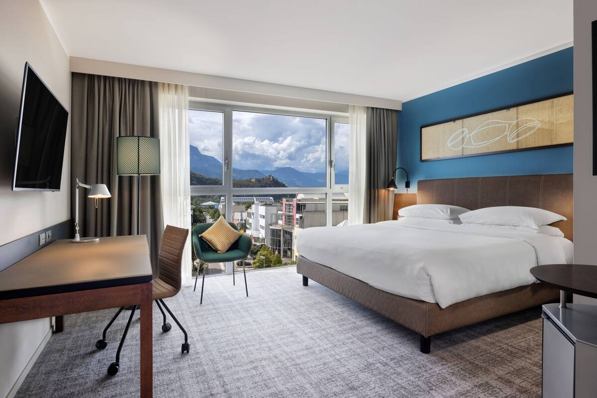 A hotel room with a bed, desk, chair, and a large window offering a city view.