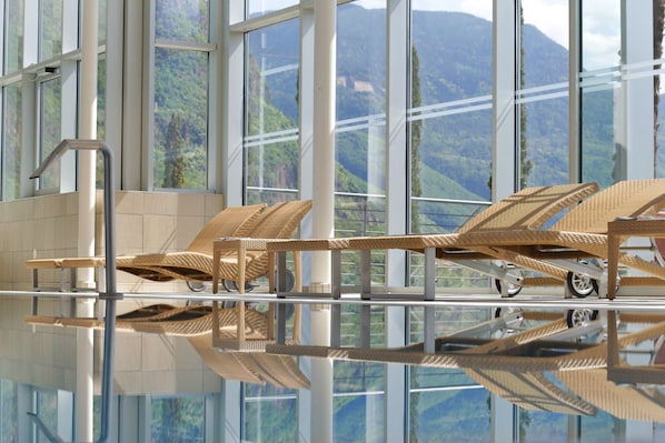 A modern indoor swimming pool with clear glass walls, offering views of a mountainous landscape.