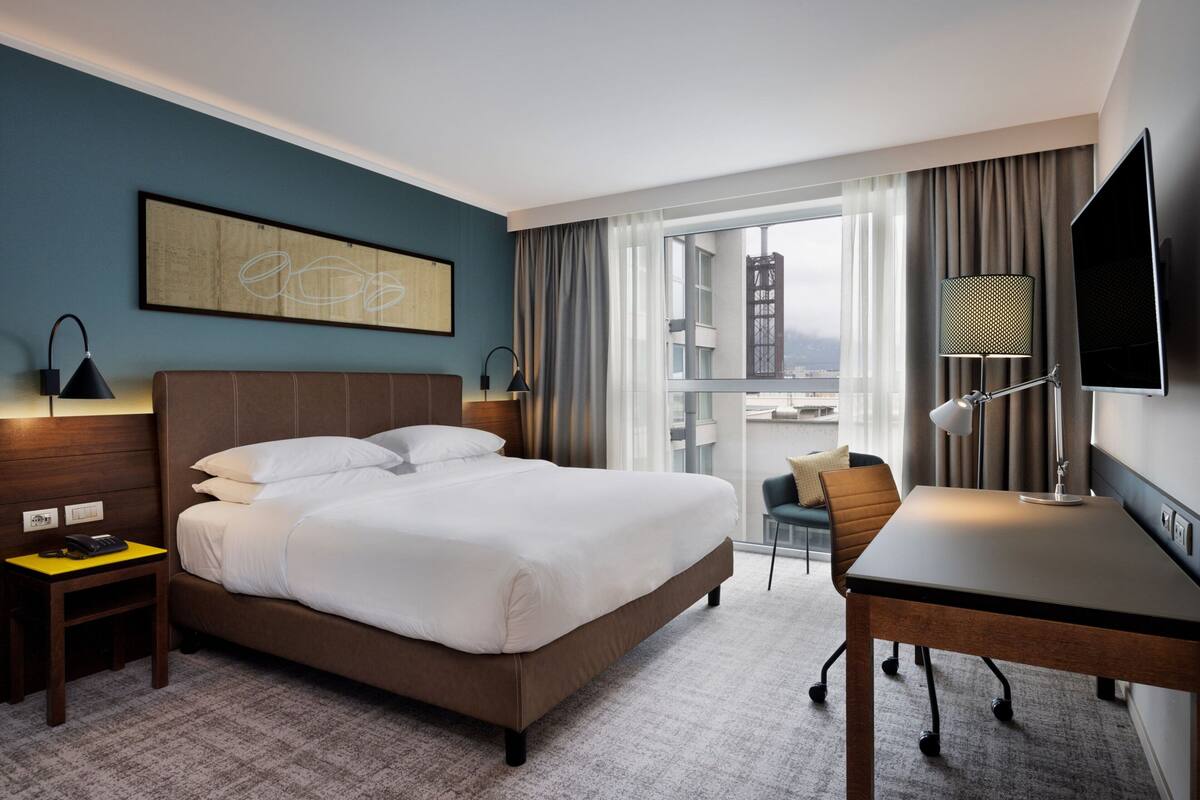 A hotel room with a large bed, a desk, a chair, a TV, and a window with curtains.