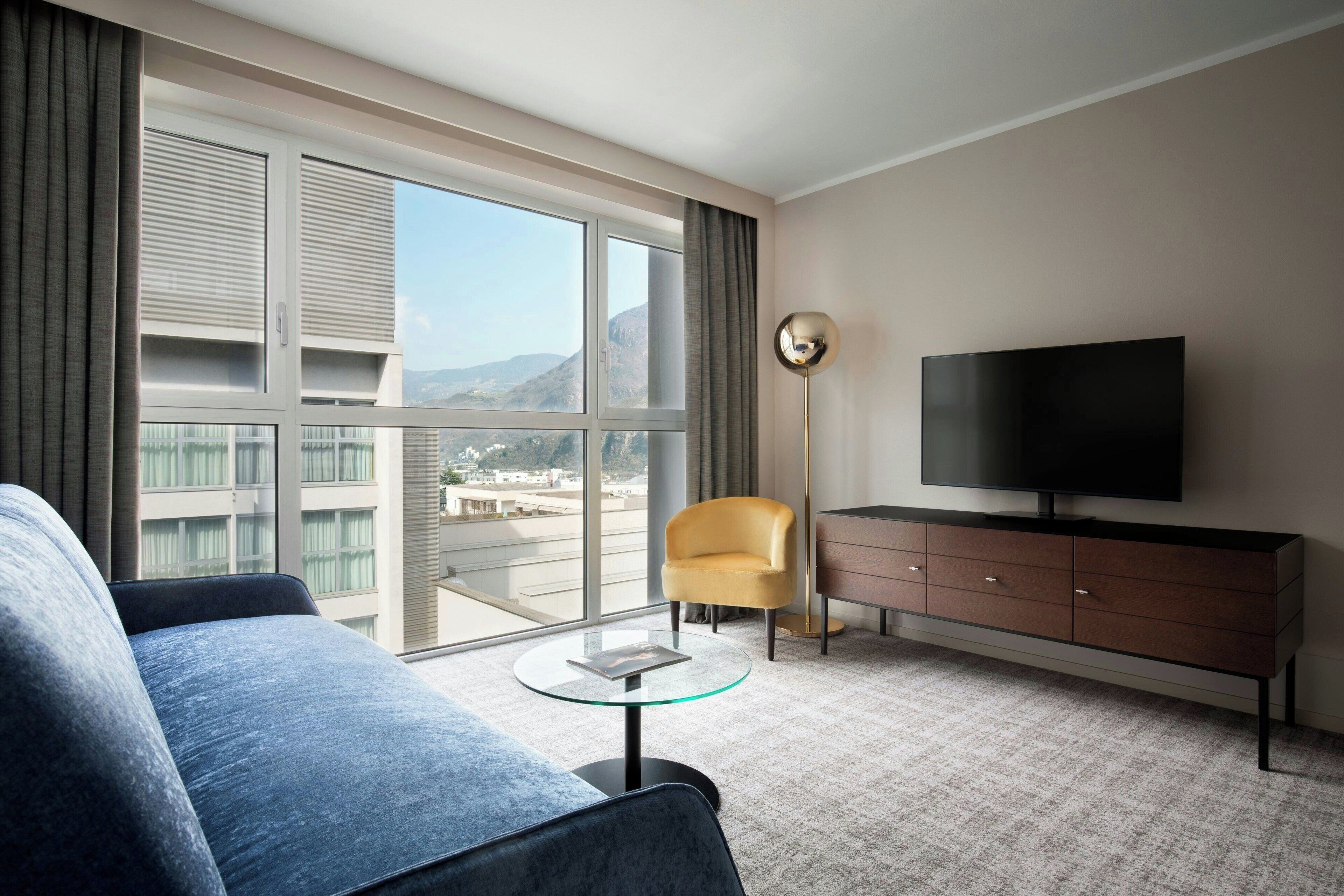 A modern hotel room with a large window, a sofa, a television, a small table, and a lamp.