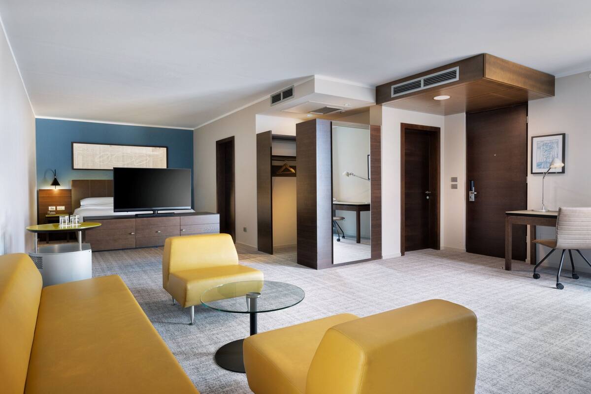 A modern hotel room with a flat-screen TV, yellow sofas, a round glass table, and a desk with a chair.