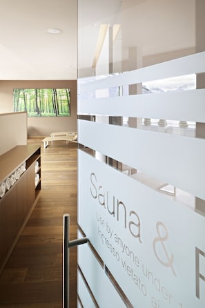 Un moderno espacio de spa con un mostrador de recepción de madera, un letrero de sauna y vistas a un bosque a través de amplios ventanales.