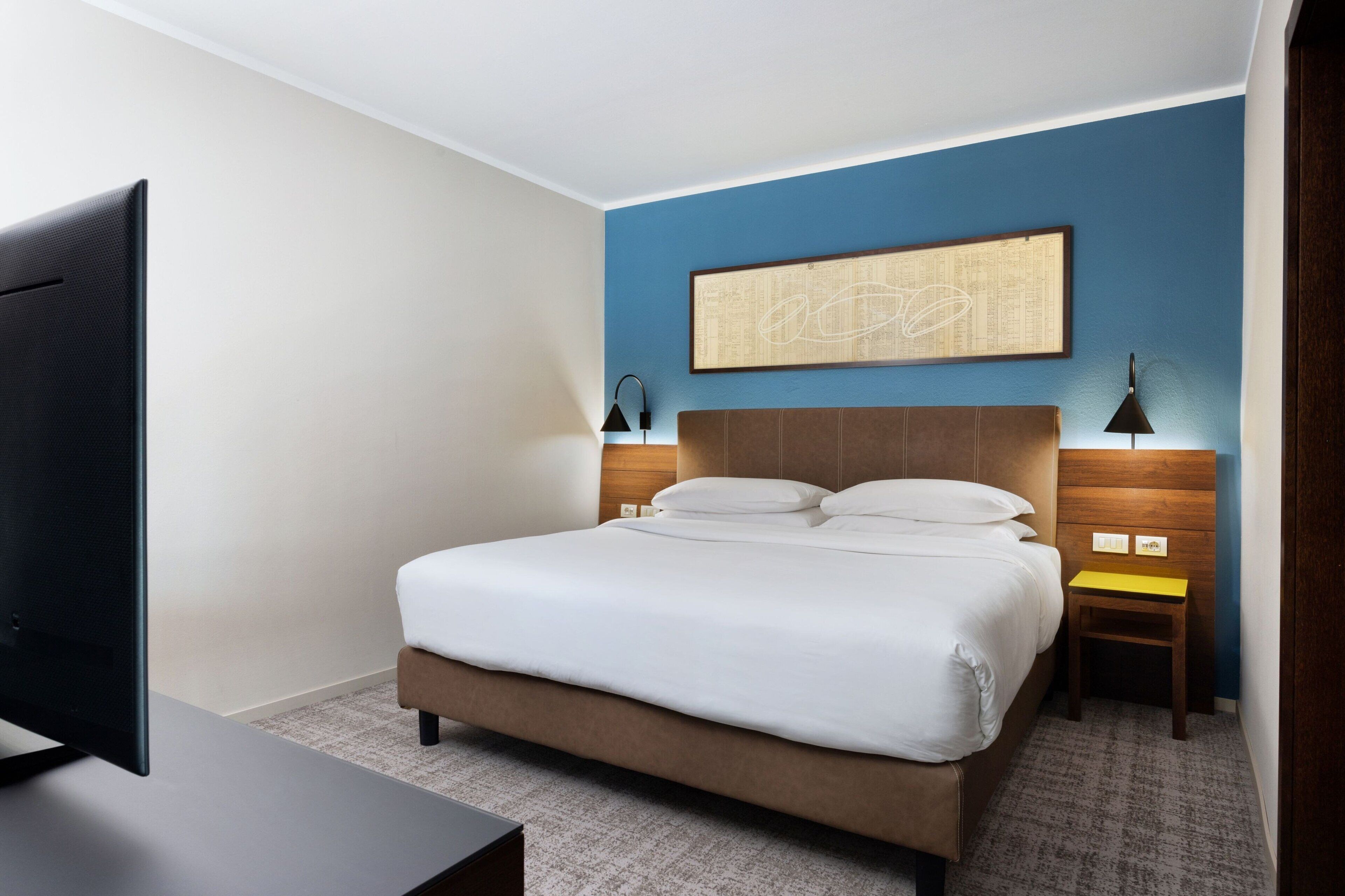 Habitación de hotel con una cama grande, un televisor de pantalla plana, cabecero de madera y una obra de arte colgada en la pared.