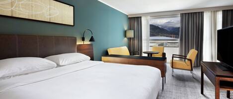 A hotel room with a large bed, a desk, a chair, and a television. There is a window with a view of mountains.