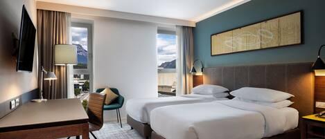 A hotel room with a bed, a desk, a chair, and a view of the mountains.