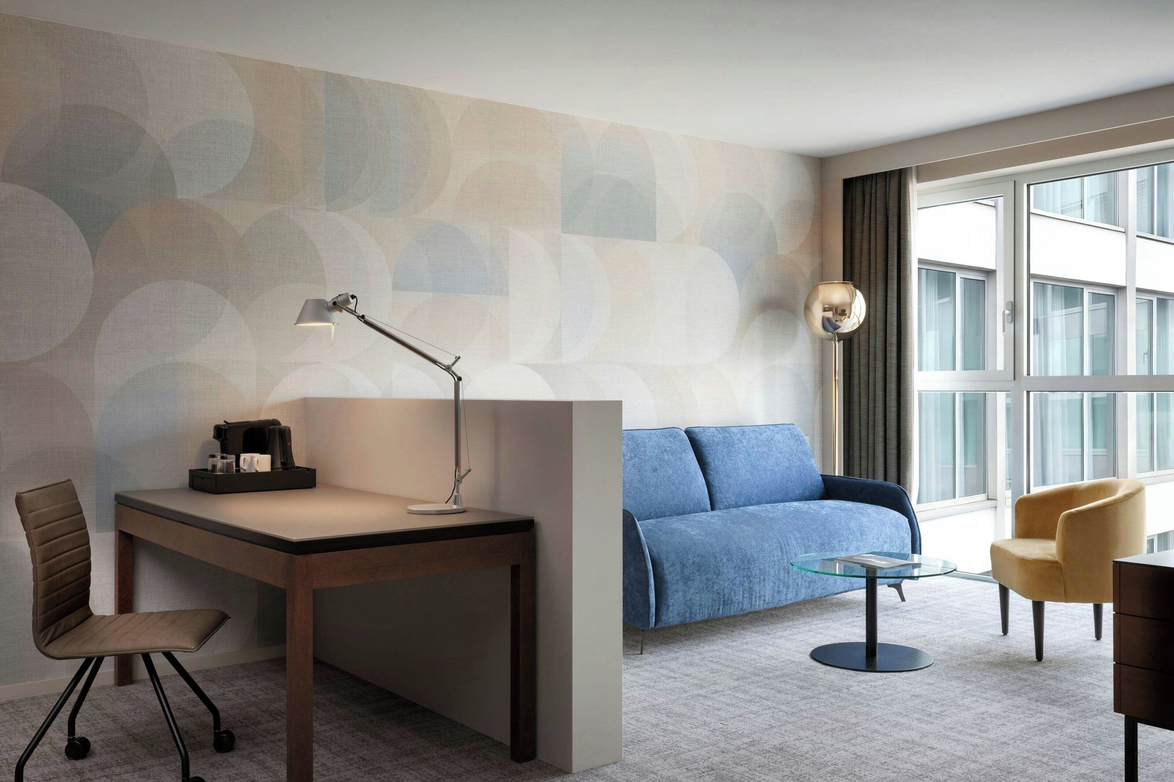 A modern hotel room with a desk, chair, sofa, and armchair, featuring a large window and abstract wall design.