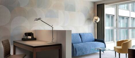 A modern hotel room with a desk, chair, sofa, and armchair, featuring a large window and abstract wall design.