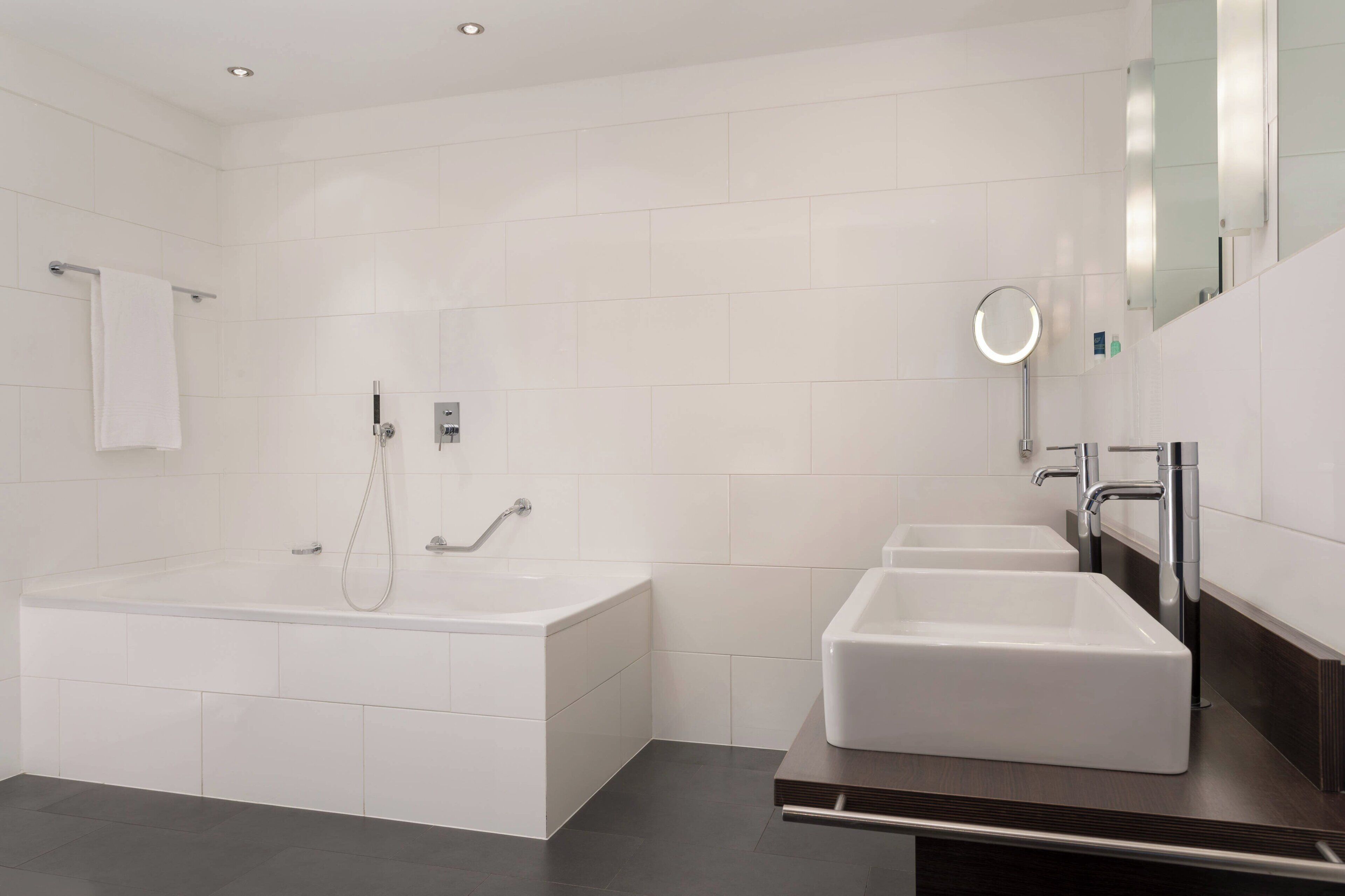 A modern bathroom with a large bathtub, two sinks, and a mirror with a light.