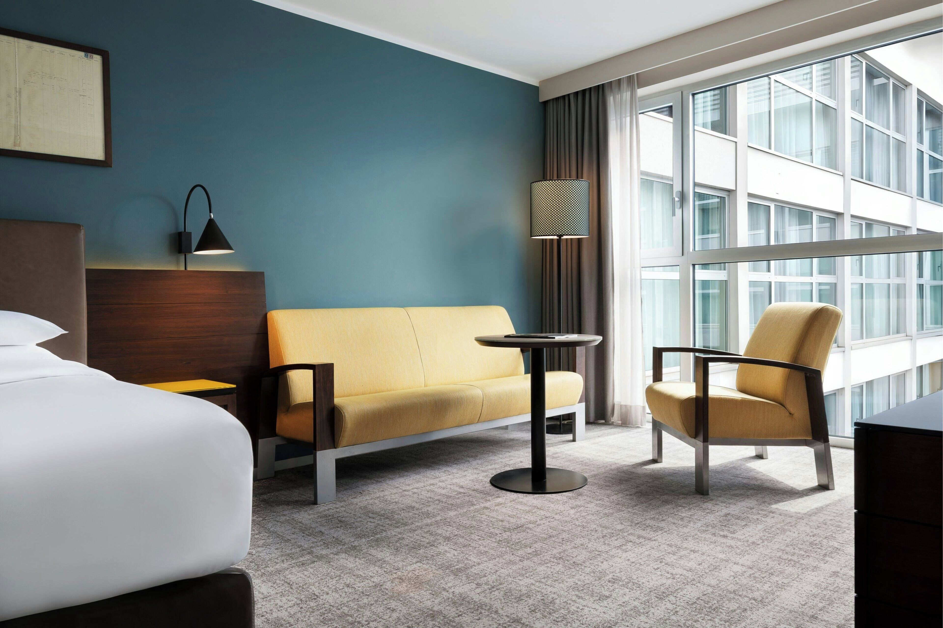 Hotelkamer met een bed, een bank, een stoel, een kleine tafel, een lamp en een groot raam met gordijnen.