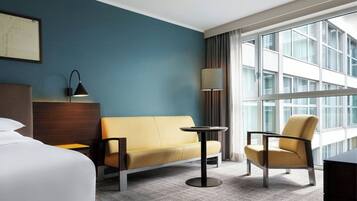 Ein Hotelzimmer mit einem Bett, einem Sofa, einem Sessel, einem kleinen Tisch, einer Lampe und einem großen Fenster mit Vorhängen.