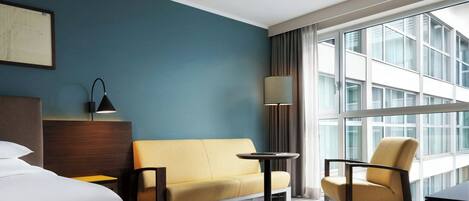Hotelkamer met een bed, een bank, een stoel, een kleine tafel, een lamp en een groot raam met gordijnen.