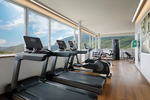 Une salle de sport équipée de tapis de course, de vélos d’exercice et d’appareils de musculation, avec de grandes fenêtres offrant une vue sur les montagnes.