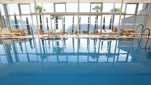 An indoor swimming pool with clear glass walls, multiple lounge chairs, and a view of mountains. - Four Points by Sheraton Bolzano (Bolzano)