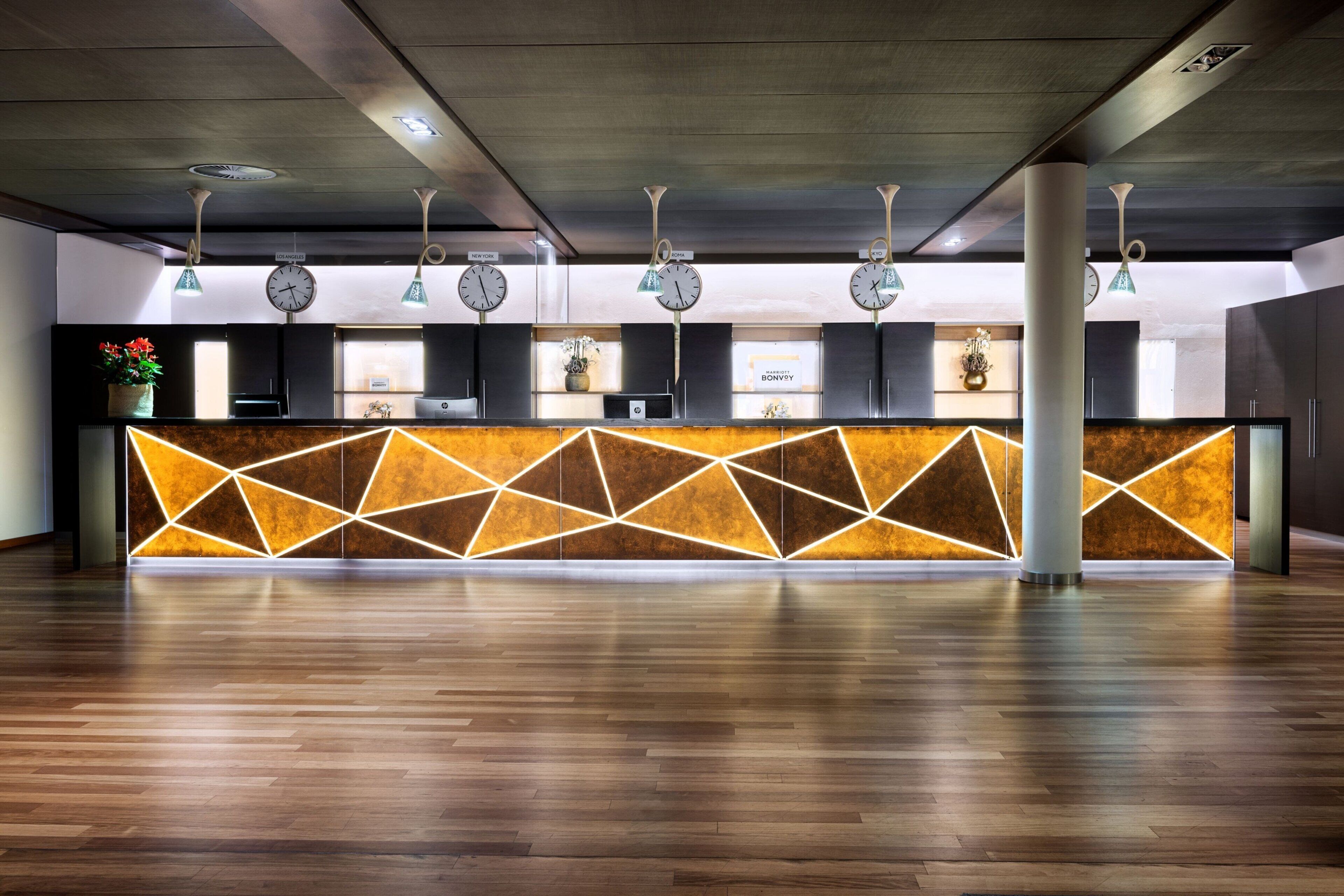 A hotel reception area with a modern design, featuring a geometric patterned wall, multiple clocks indicating different time zones, and a clean, polished wooden floor.