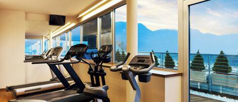 Ein Fitnessraum mit Laufbändern und Crosstrainern, großen Fenstern mit Blick auf Berge und einer Holzterrasse draußen.