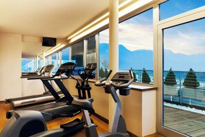 Un gimnasio con cintas de correr y elípticas, amplias ventanas con vistas a la montaña, y una terraza de madera en el exterior.