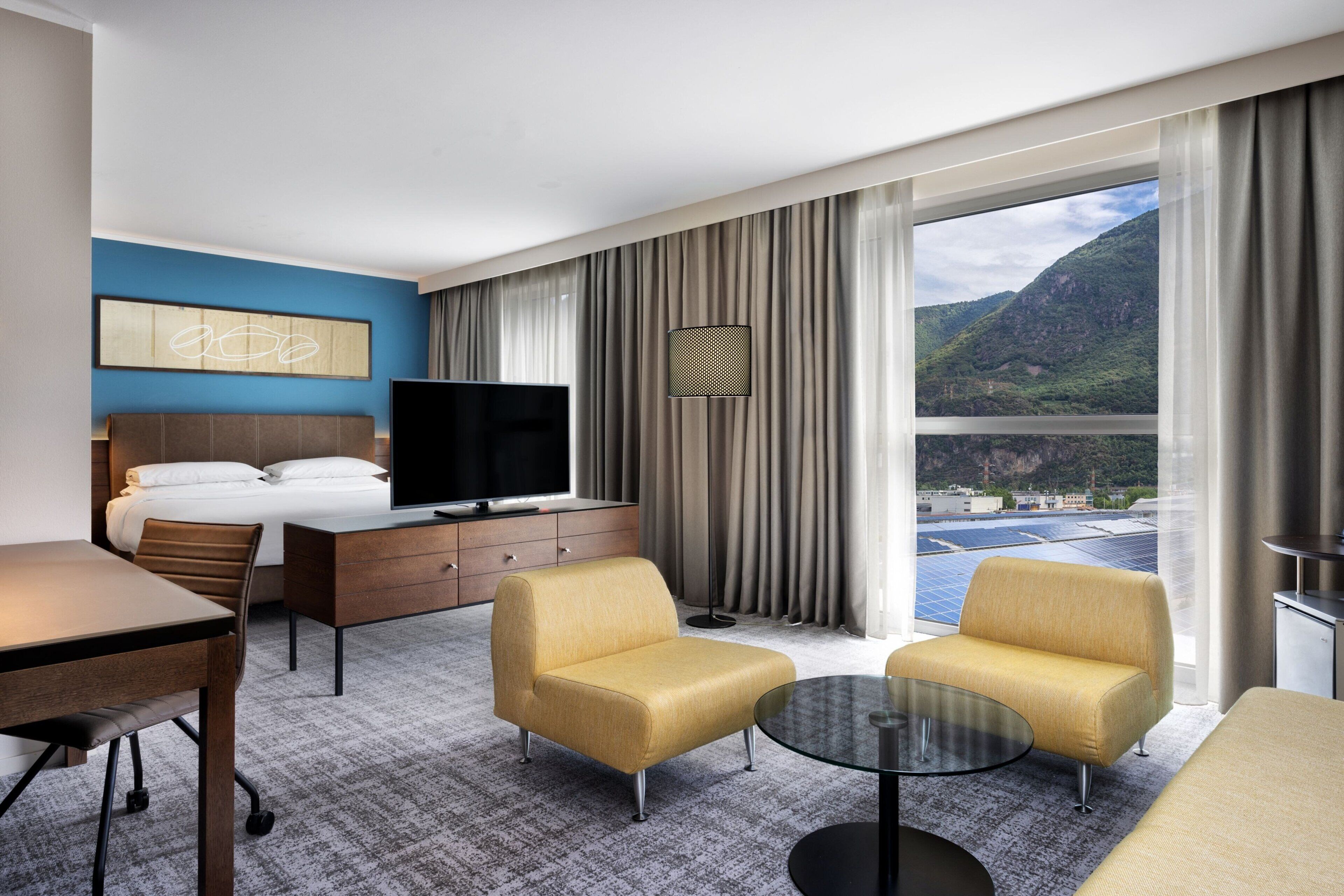 Ein Hotelzimmer mit Bett, Fernseher, Schreibtisch, zwei Sesseln, einem kleinen Tisch und Blick auf eine gebirgige Landschaft durch große Fenster.