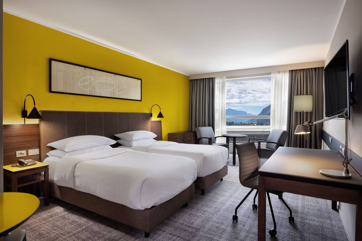 A hotel room with two beds, a desk, a chair, a TV, and a window with a view of mountains.