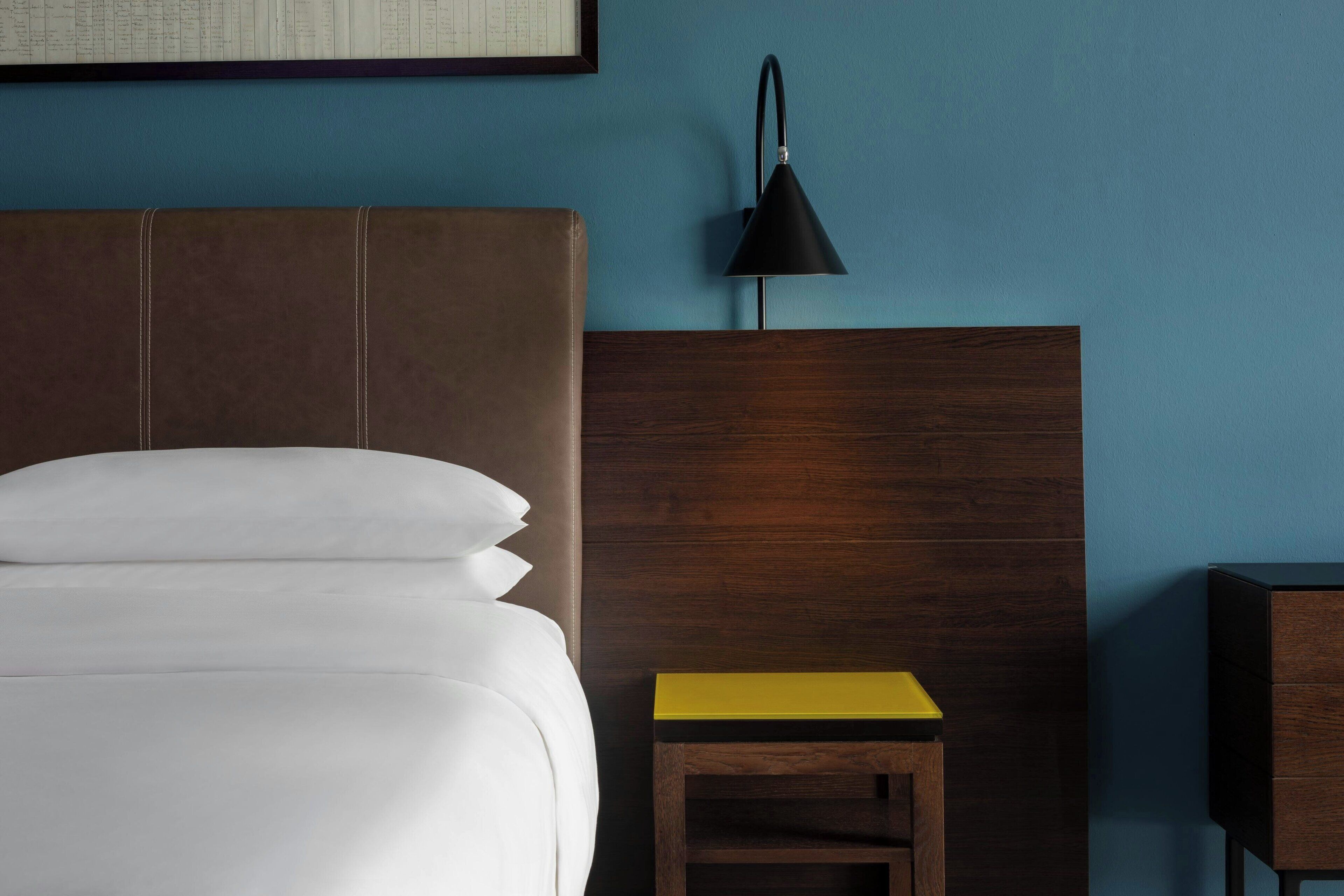 A bed with white linens, a wooden headboard, a small wooden nightstand with a yellow top, and a black desk lamp.