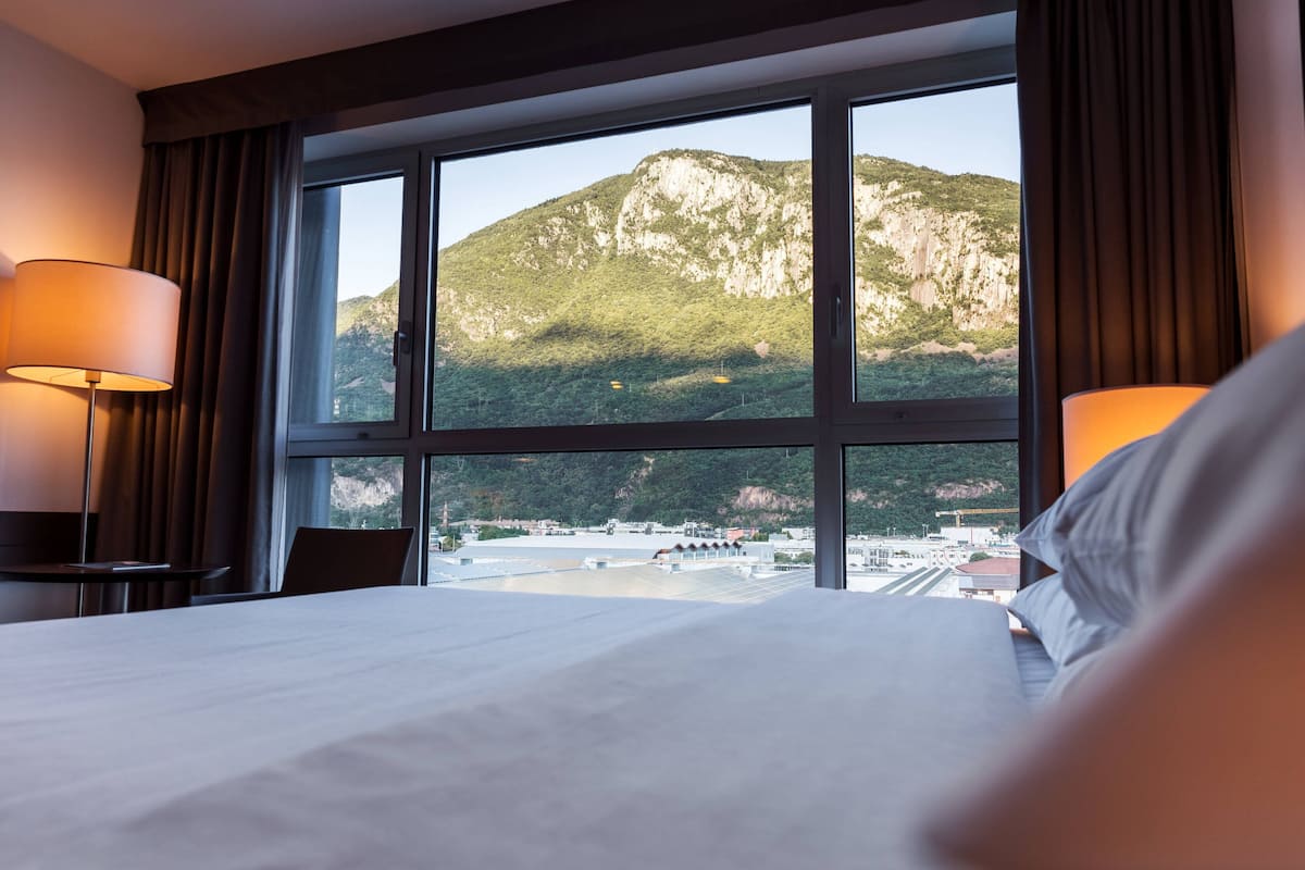 Habitación de hotel con una cama grande, dos lámparas de noche y vistas a un paisaje montañoso a través de un ventanal amplio.