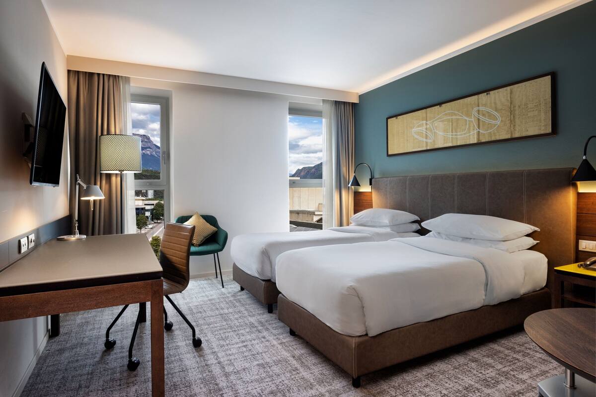 A hotel room with two beds, a desk, a chair, a TV, and a view of mountains.