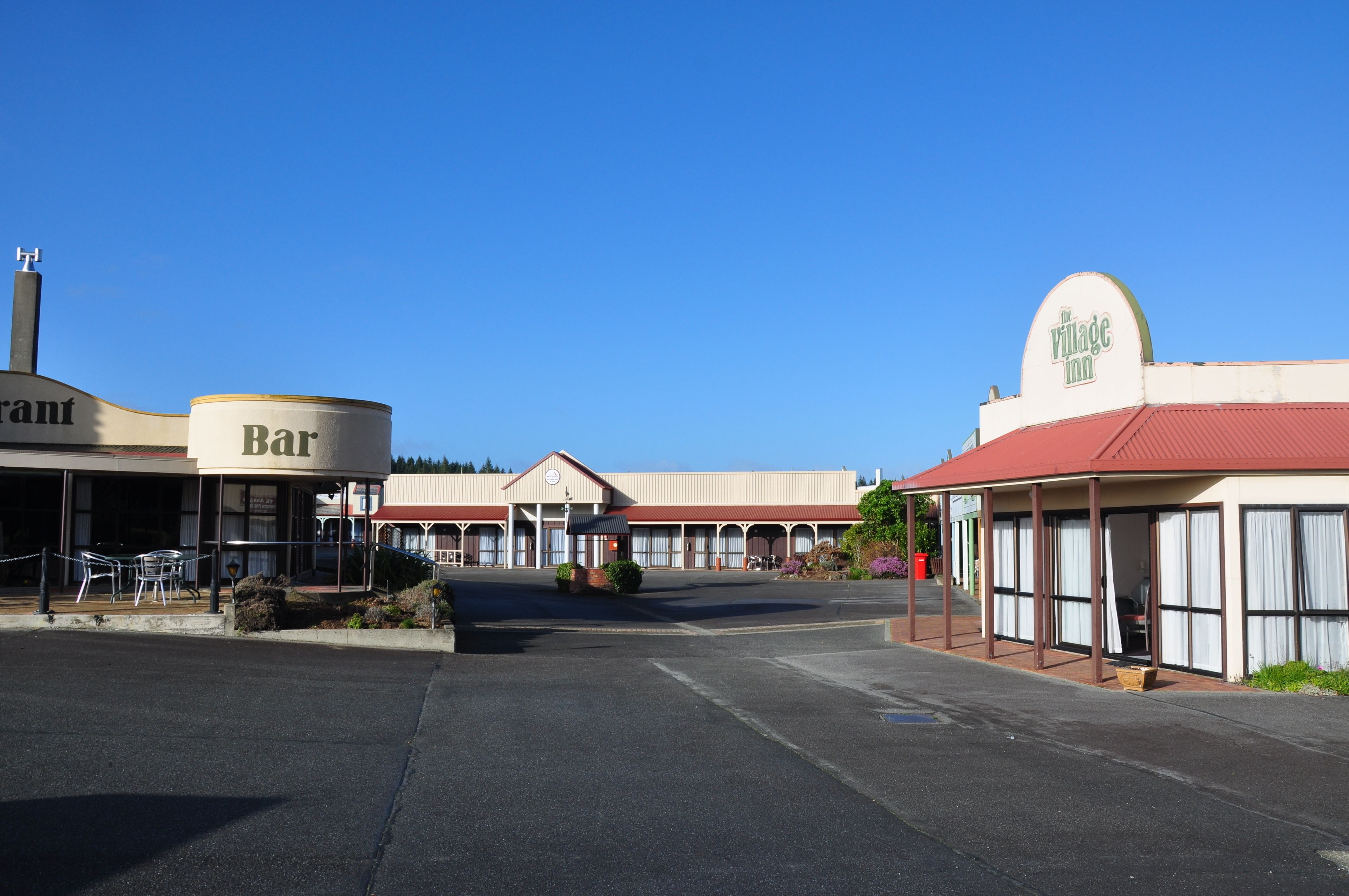 Un edificio con un letrero que dice 'Bar' y otro que dice 'Village Inn', con un área de asientos al aire libre cubierta y un estacionamiento.