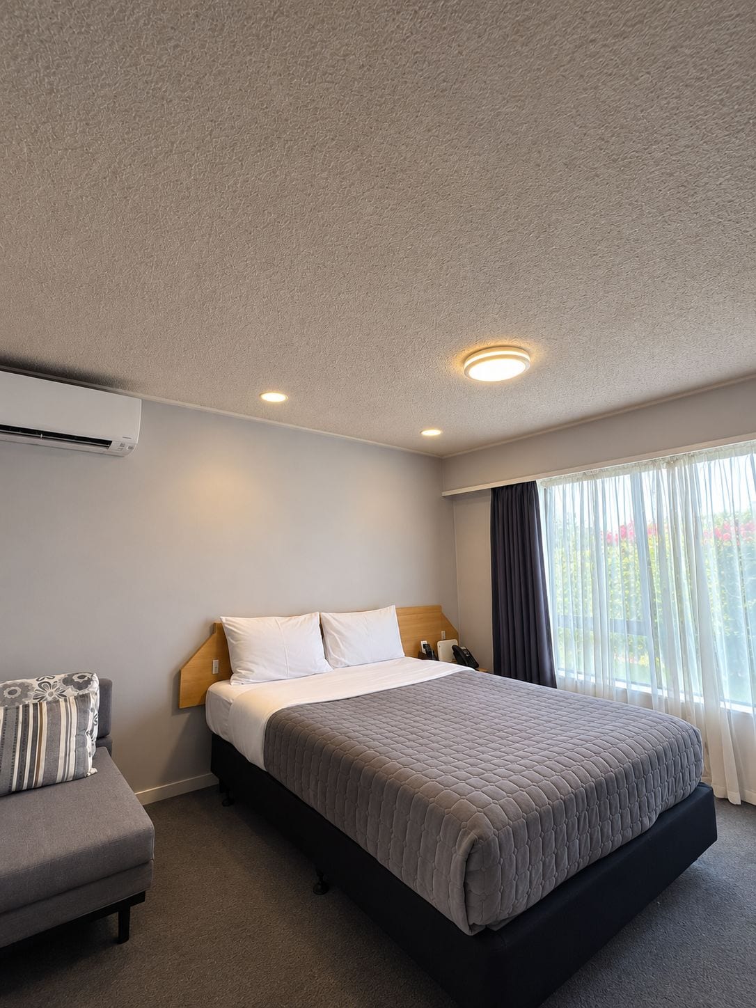 A hotel room with a bed, bedside table, and an air conditioning unit.