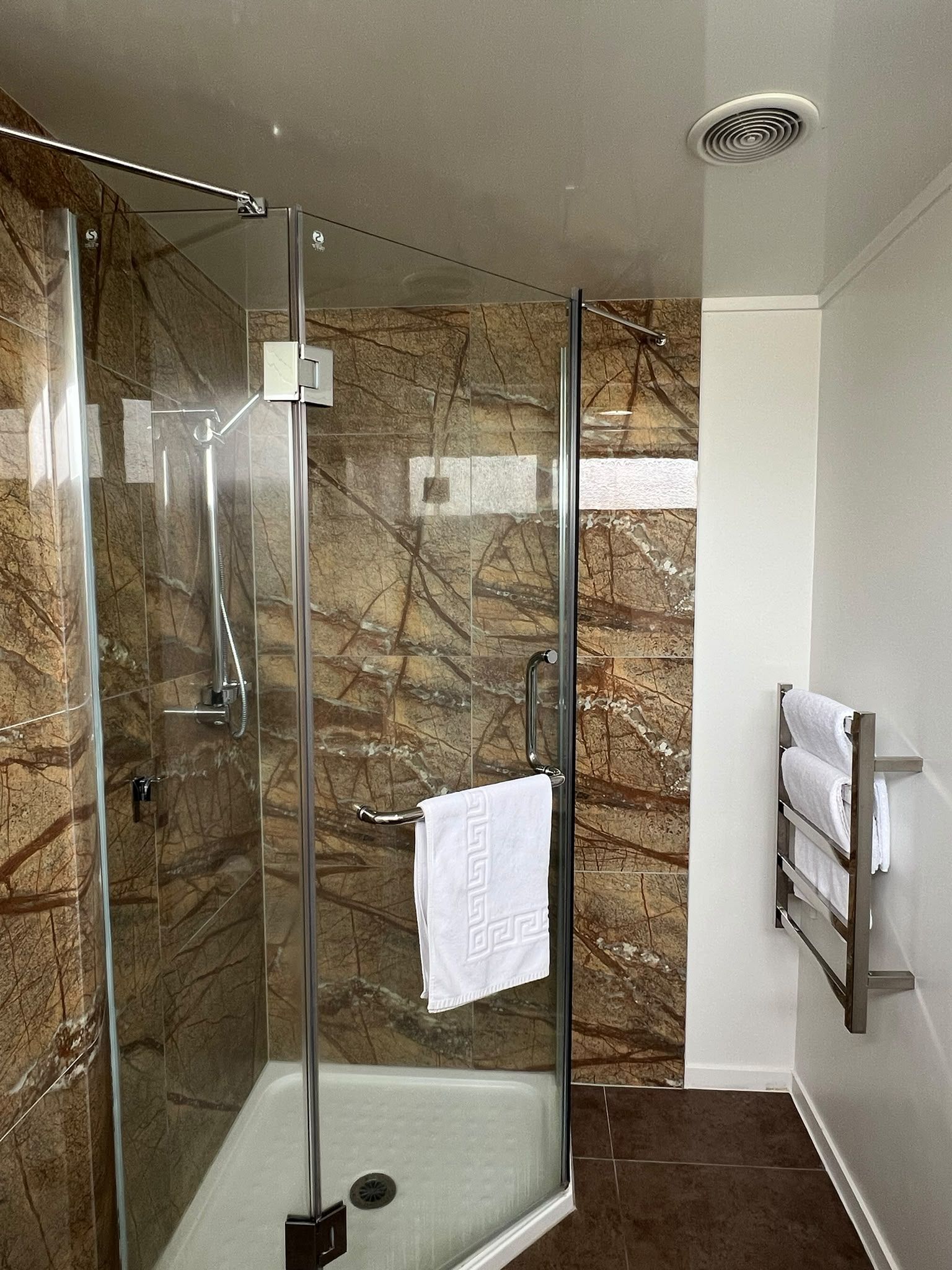 A glass-enclosed shower with a marble wall backdrop, a towel rack, and a vent on the ceiling.