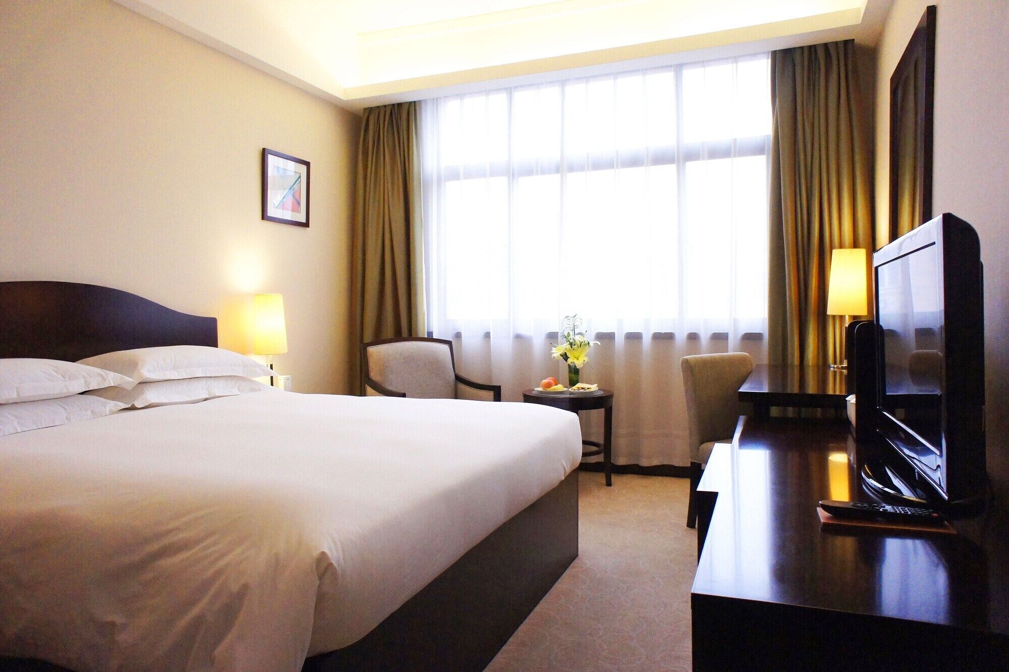 A hotel room with a large bed, a desk with a television, a chair, and a small table with a vase of flowers.