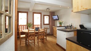 A kitchen with wooden cabinets, a dining table with chairs, a television mounted above the counter, and a window with curtains. - Résidence Val Claret (Tignes)