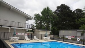 A rectangular pool with a blue interior, surrounded by a black metal fence. There are lounge chairs and a table with a table umbrella. A building with a balcony and multiple signs is visible in the background. - Americas Best Value Inn Goldsboro (Goldsboro)