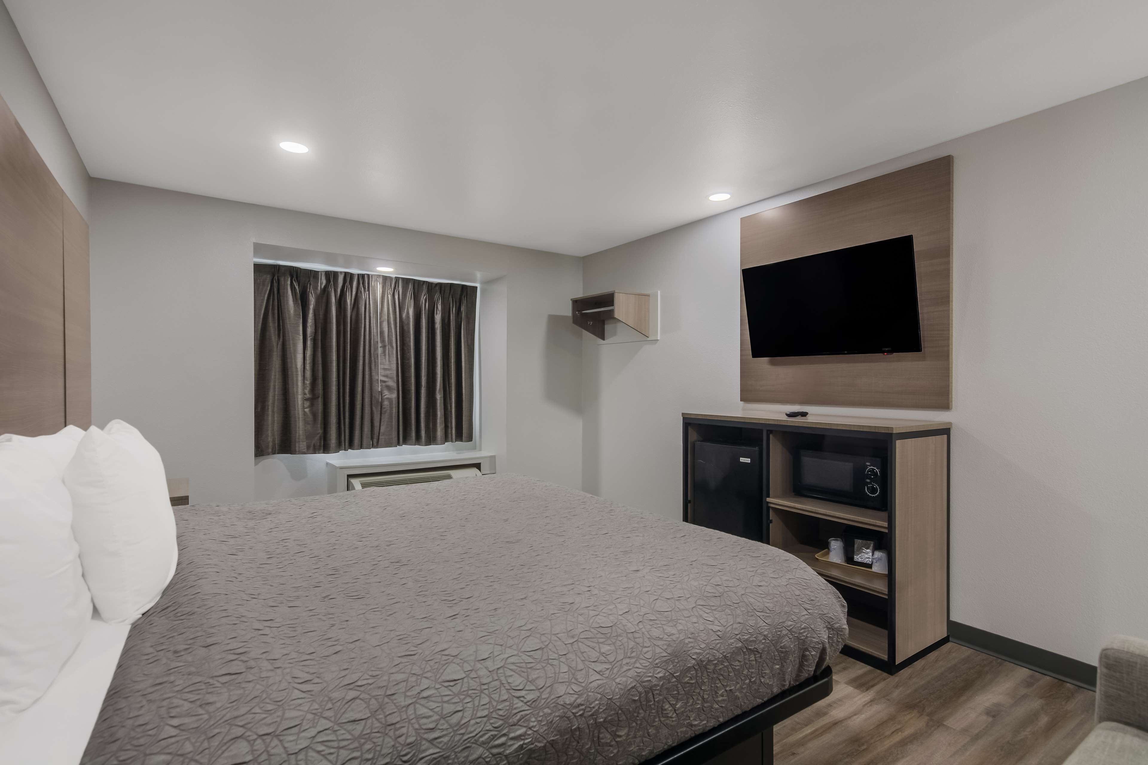 A modern hotel room with a large bed, a flat-screen TV mounted on the wall, a built-in TV stand with a microwave, and a window with curtains.