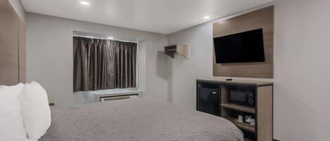 A modern hotel room with a large bed, a flat-screen TV mounted on the wall, a built-in TV stand with a microwave, and a window with curtains.