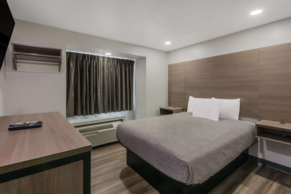 A modern hotel room with a bed, a desk, a window with curtains, a bedside table, and a wall-mounted air conditioning unit.