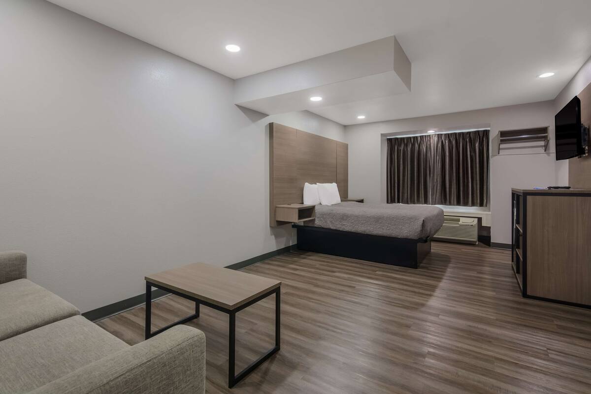 A modern hotel room with a bed, a small table, a sofa, a TV, and a window with curtains.