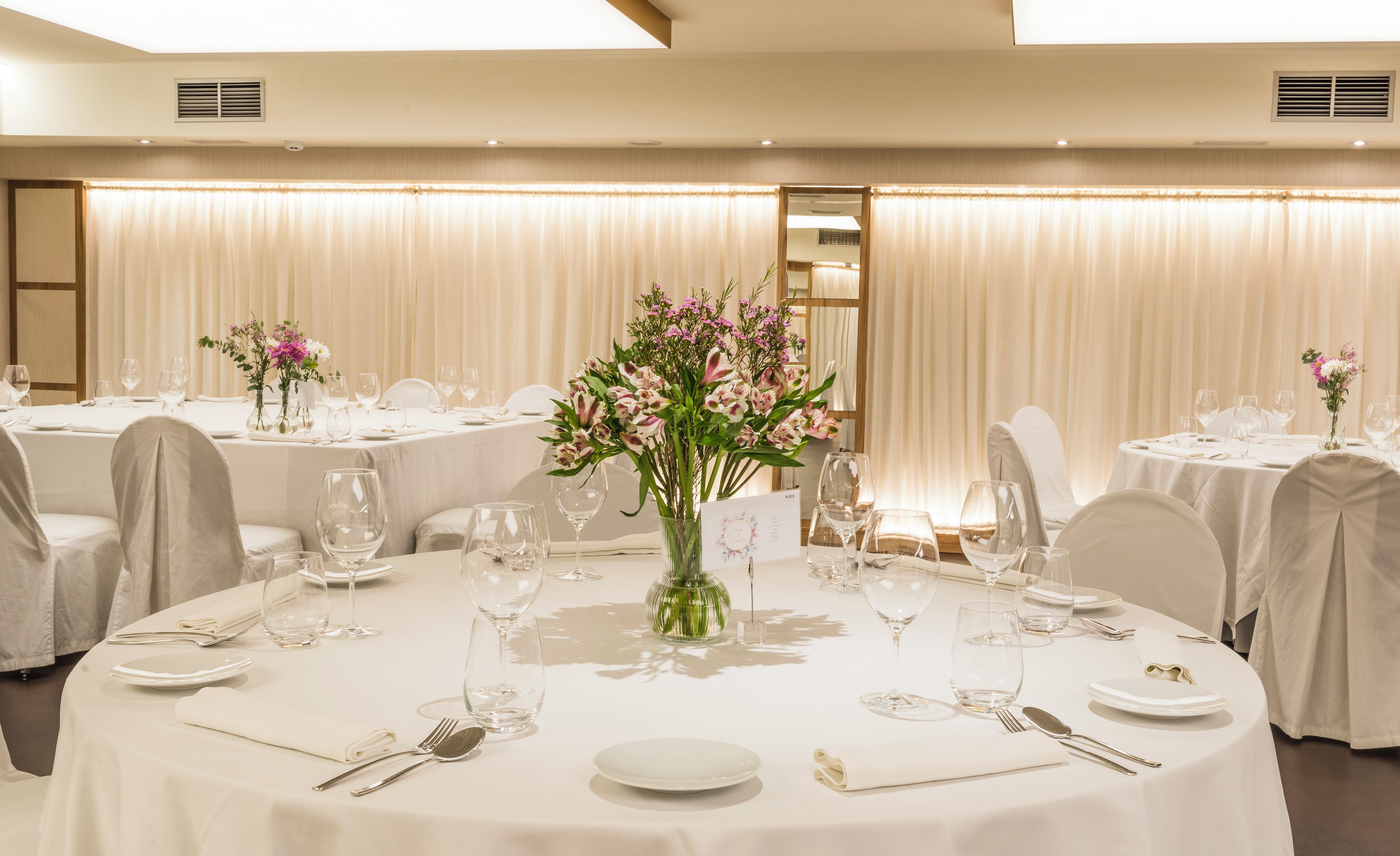 Un salón de banquetes con mesas redondas dispuestas para el servicio de comidas, cada una con platos, cubiertos y copas de vino.