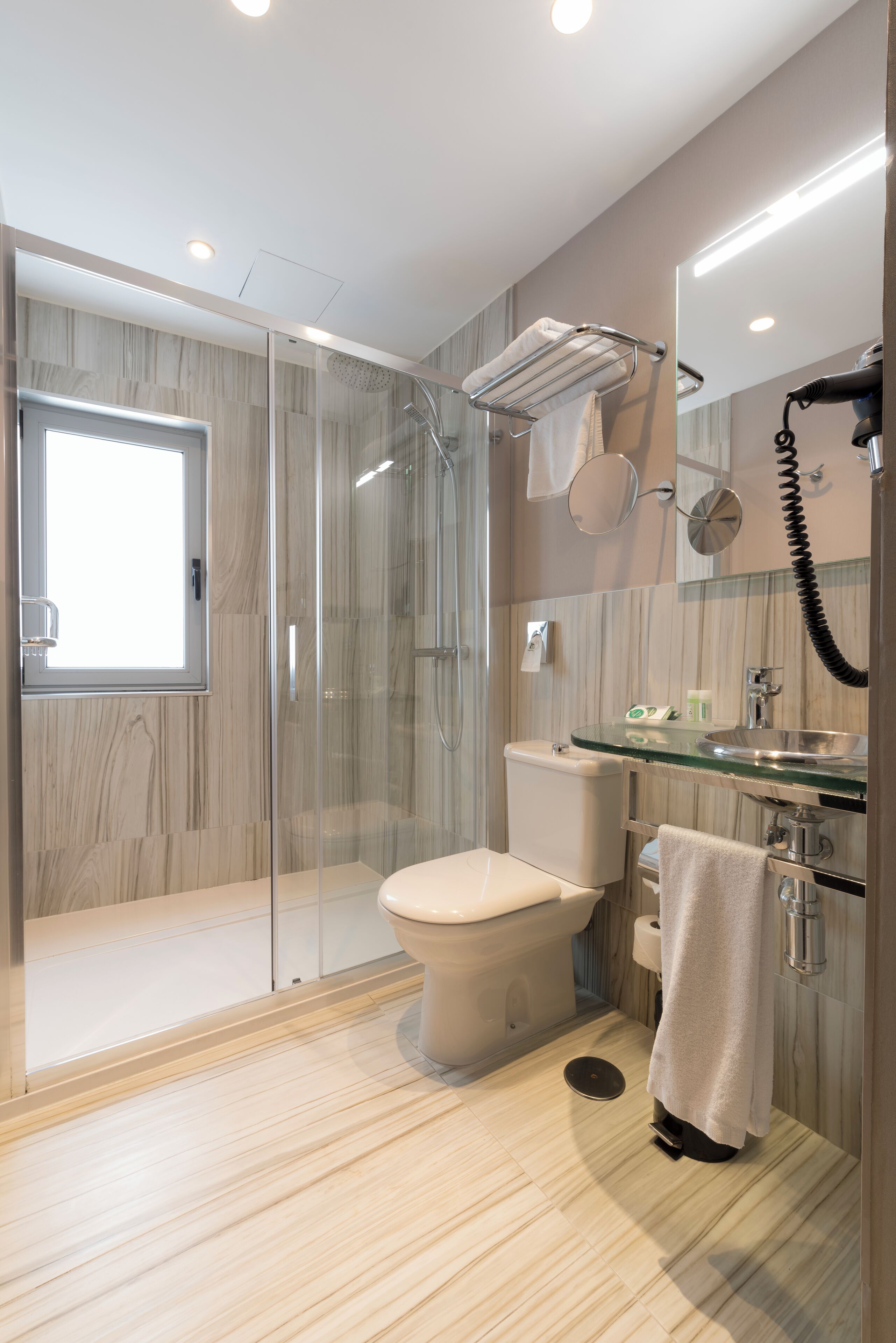 Un baño moderno con cabina de ducha de vidrio, inodoro, lavamanos con encimera de vidrio y un perchero para toallas.
